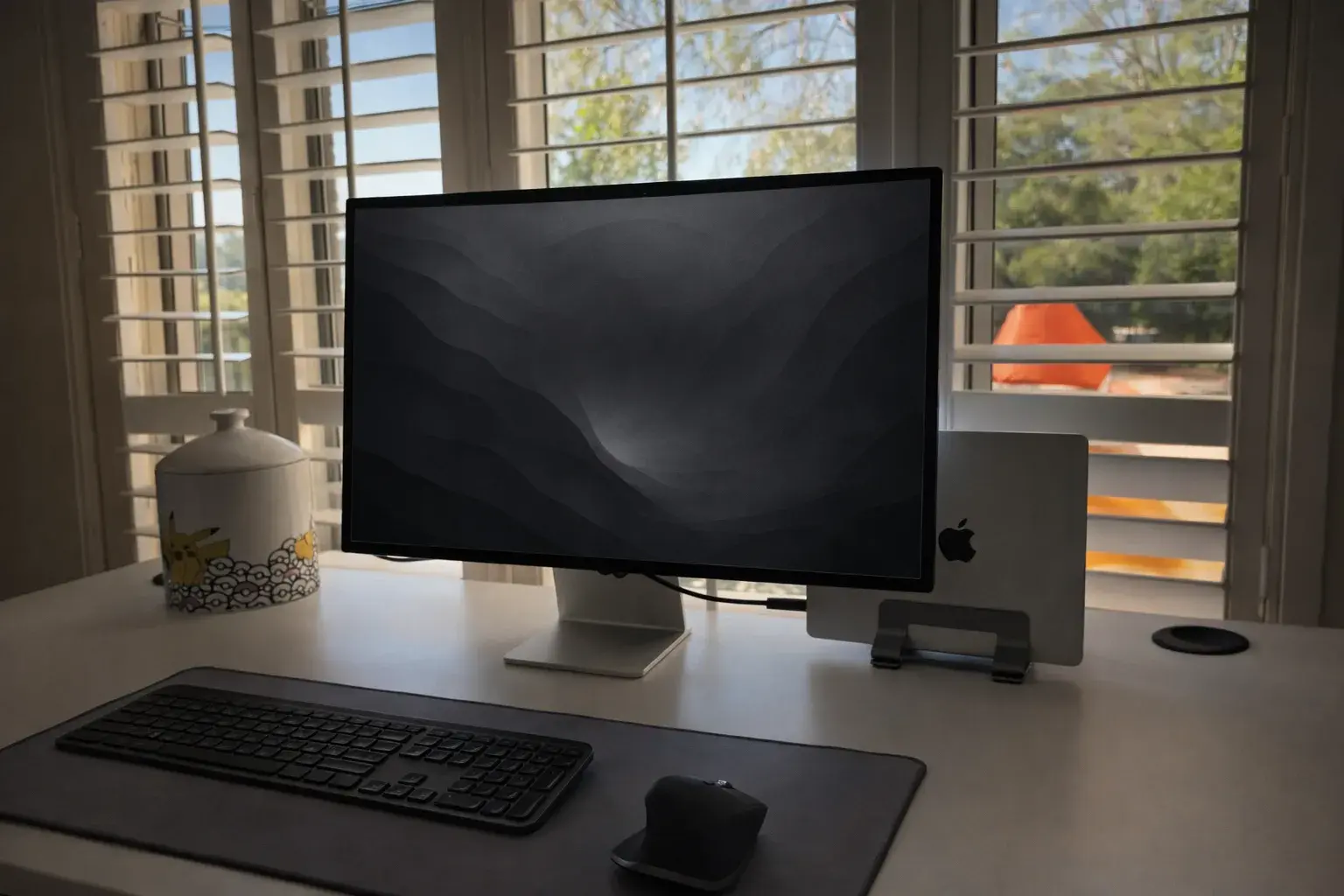 Workspace with a monitor, keyboard, mouse, and laptop on a white desk in front of bright window shutters